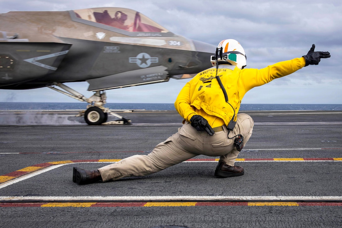 A sailor leans to the right while signaling to an aircraft as it launches from a ship at sea on a gloomy day.