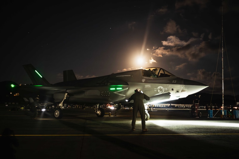 A Marine shines a flashlight next to an aircraft in the dark illuminated by white and green lights.