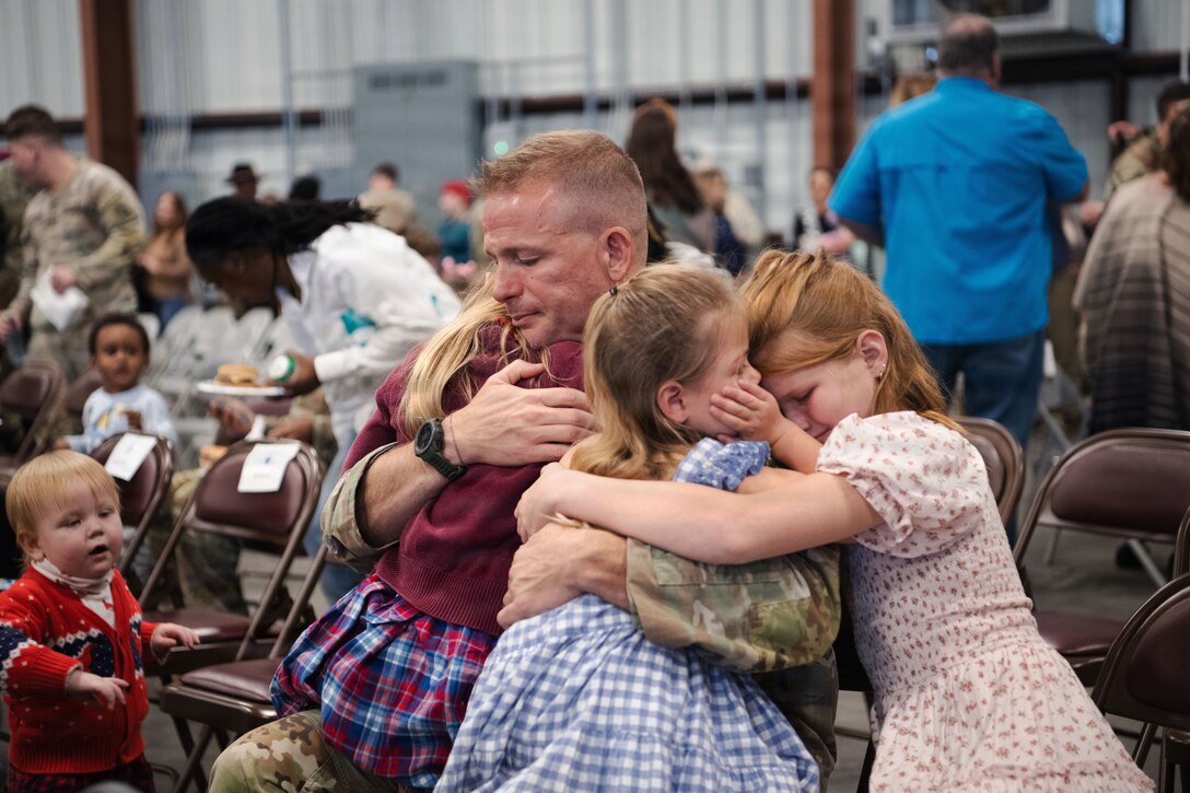 A man tightly hugs three children in a crowded room following a ceremony.