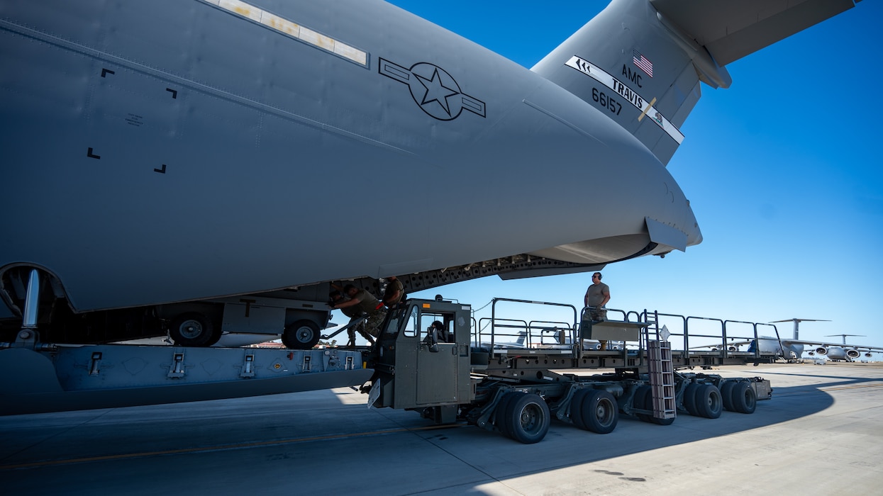 The tail of an aircraft is visible while being loaded by Airmen on a flight line