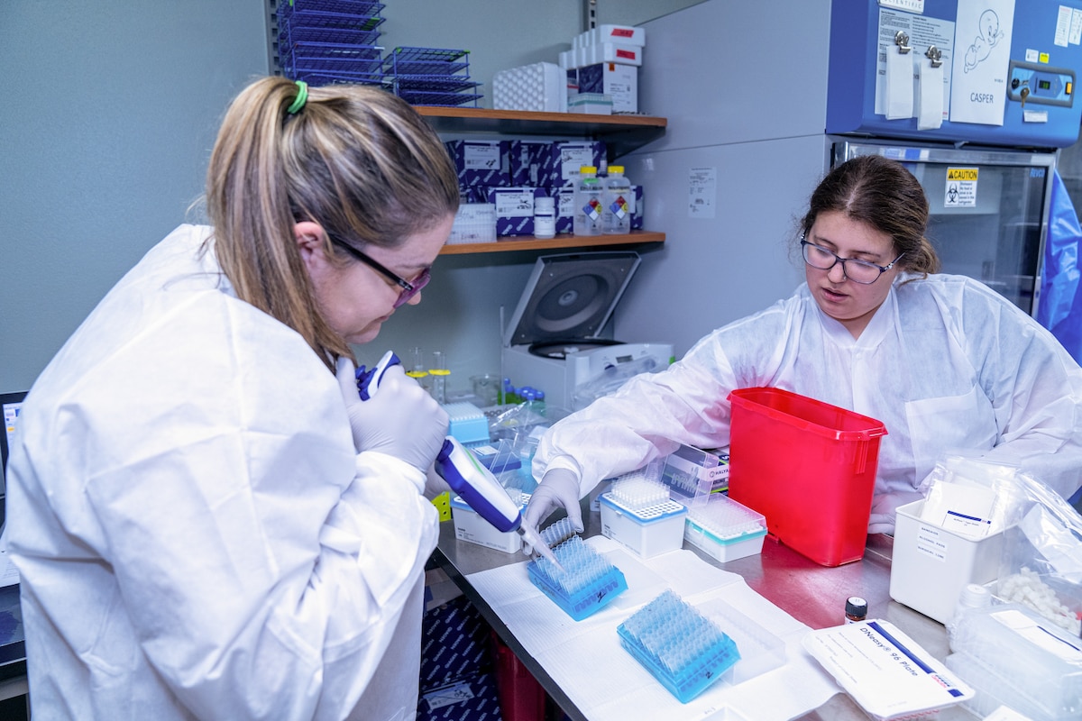 Two women, one blonde and the other brunette, in white lab coats wearing blue latex gloves handle blood samples sitting on a table.