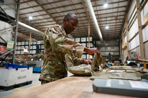 An Airman in a warehouse opens flaps of a tactical vest lying on a table