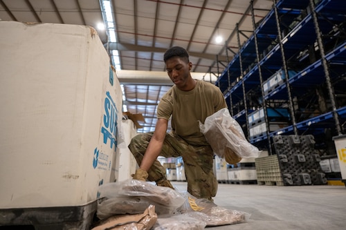 An Airman in a warehouse kneels next to a cardboard bin sorting items lying on the ground