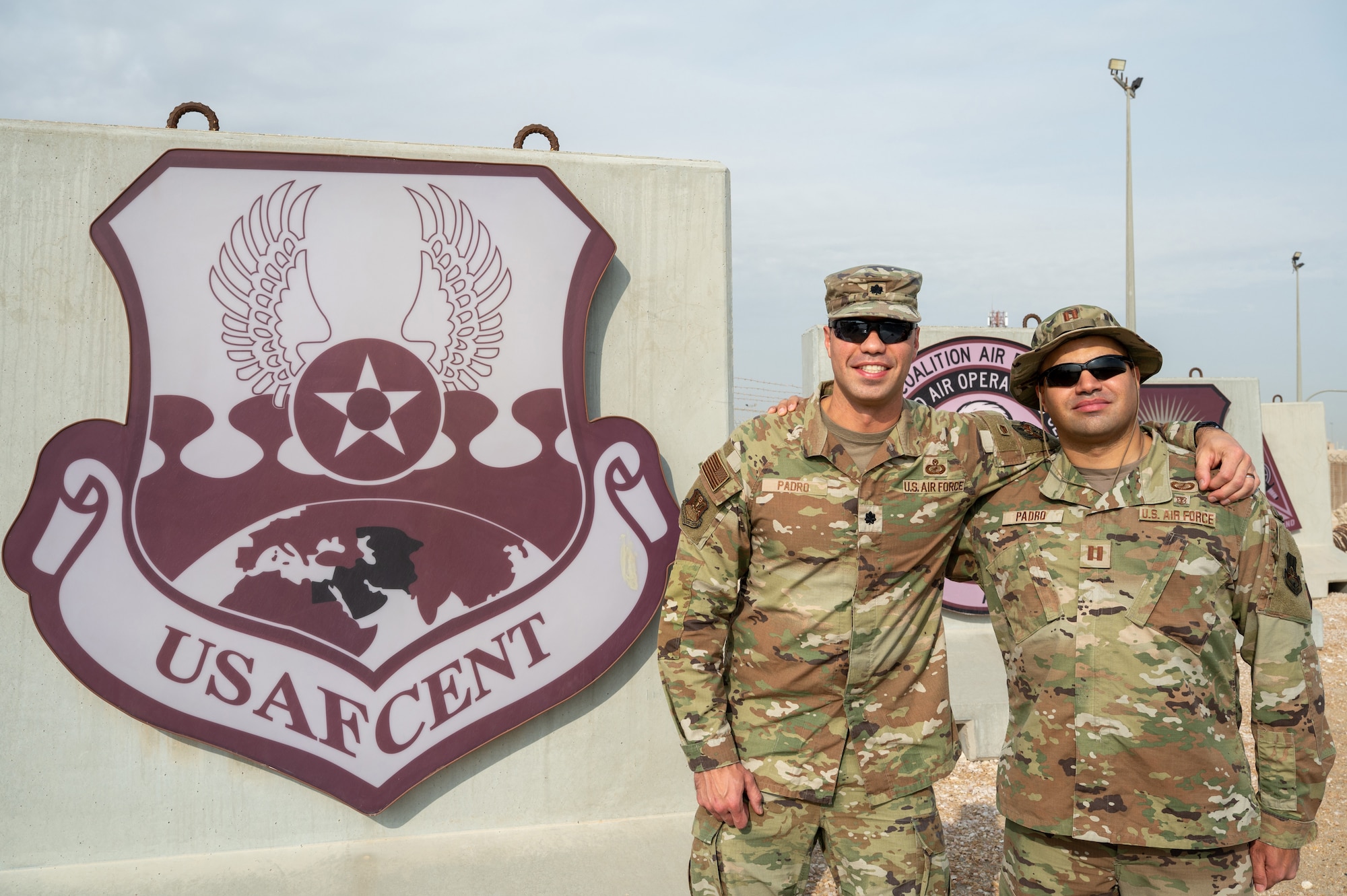Two uniformed members stand side-by-side next to the U.S. Air Forces Central insignia