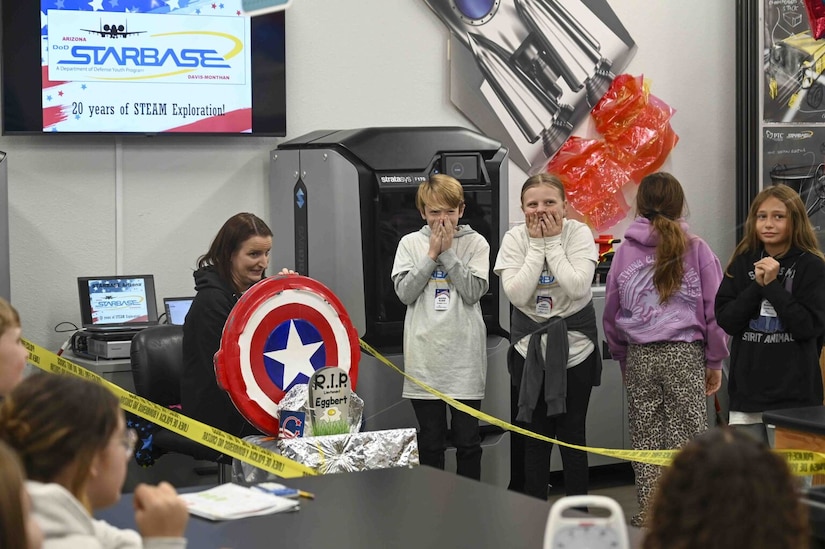 Five children stand before a group of seated children. One child, a girl, holds a model Captain America shield. Two children hold their hands over their mouths as one other has a fearful look on her face and the other faces away from the group.