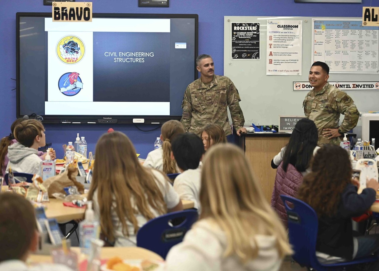 Two military members stand at the front of a classroom to instruct a group of seated children.