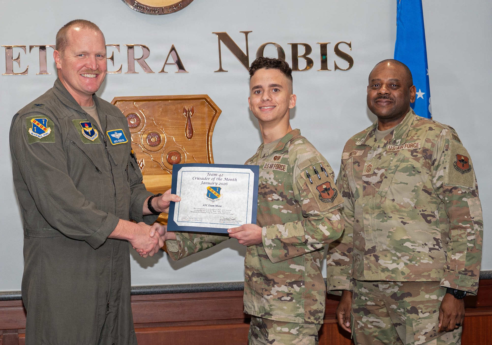 U.S Air Force Col. Adam Rudolphi, 42d Air Base Wing deputy commander, and Chief Master Sgt. Demetrius Booth, 42d ABW command chief, present the Crusader of the Month award for January to Airman 1st Class Liam Mora, military justice paralegal, 42d ABW Legal Office, at Maxwell Air Force base, Ala. Dec. 11, 2025. This recognition honors exceptional individuals who play a vital role in advancing Team Maxwell’s mission, vision and priorities. (U.S. Air Force photo by Senior Airman Elizabeth Figueroa)