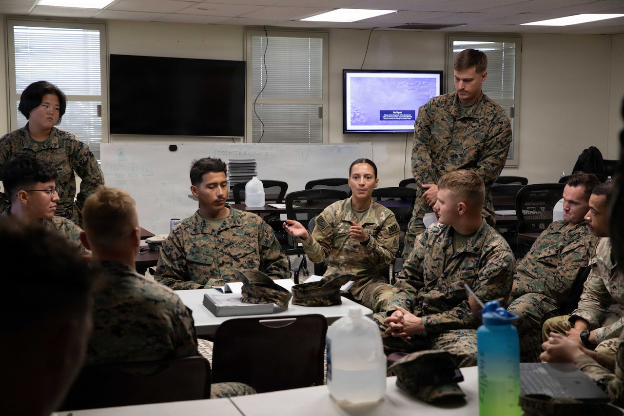 A group of people in camouflage military uniforms sit and stand around a small classroom table.
