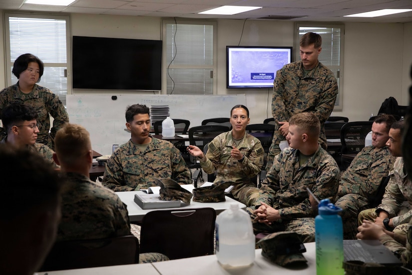 A group of people in camouflage military uniforms sit and stand around a small classroom table.