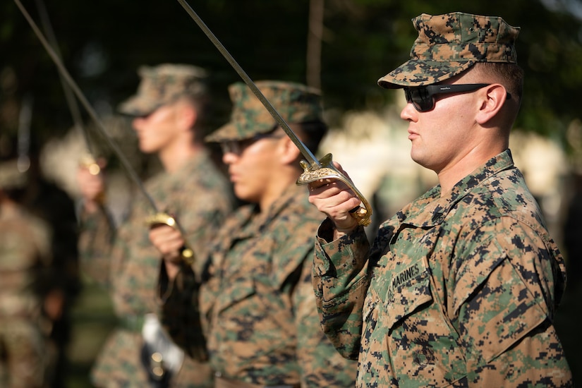 Three people in camouflage military uniforms and sunglasses hold swords while standing in formation outside.