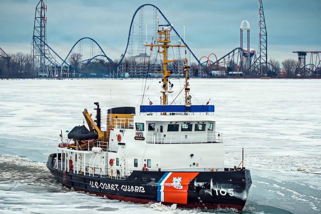 A large ship, with “U.S. Coast Guard” written on the side, moves through frozen water with an amusement park in the background.