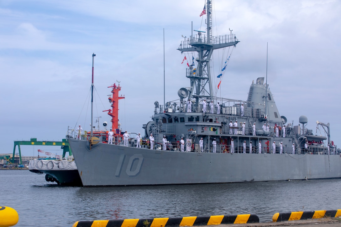 A smaller Navy ship with the crew standing along the rails in white uniforms.