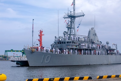 A smaller Navy ship with the crew standing along the rails in white uniforms.