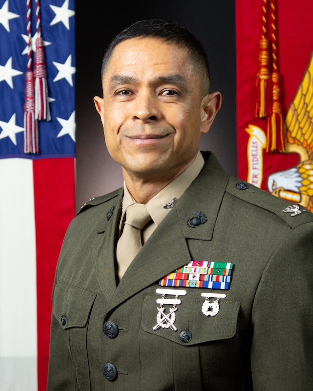 Photograph of Colonel Rico Acosta in the Service Alpha Uniform (Olive drab suitcoat over a khaki long-sleeve shirt, khaki tie) in front of flags. The American flag is on the left. The Marine Corps flag is on the right.  Colonel rank insignia appears on either side of the collar of the khaki shirt and large rank insignia appears on the epaulettes of the olive drab coat. On the left breast (right side from the viewer's point of view) are awards, including the Meritorious Service Medal with Gold Star, Navy Commendation Medal, Navy Achievement Medal with Gold Star, Joint Meritorious Unit Award, Navy Unit Commendation with Small Bronze Star, National Defense Service Medal, Afghanistan Campaign Medal with Small Bronze Star, Global War on Terrorism Expeditionary Service Medal, Global War on Terrorism Service Medal, Sea Service Deployment Ribbon with Three Small Bronze Stars, and the NATO Medal. Col Acosta is also wearing an Expert Rifle badge with retaining bar and an Expert Pistol badge.