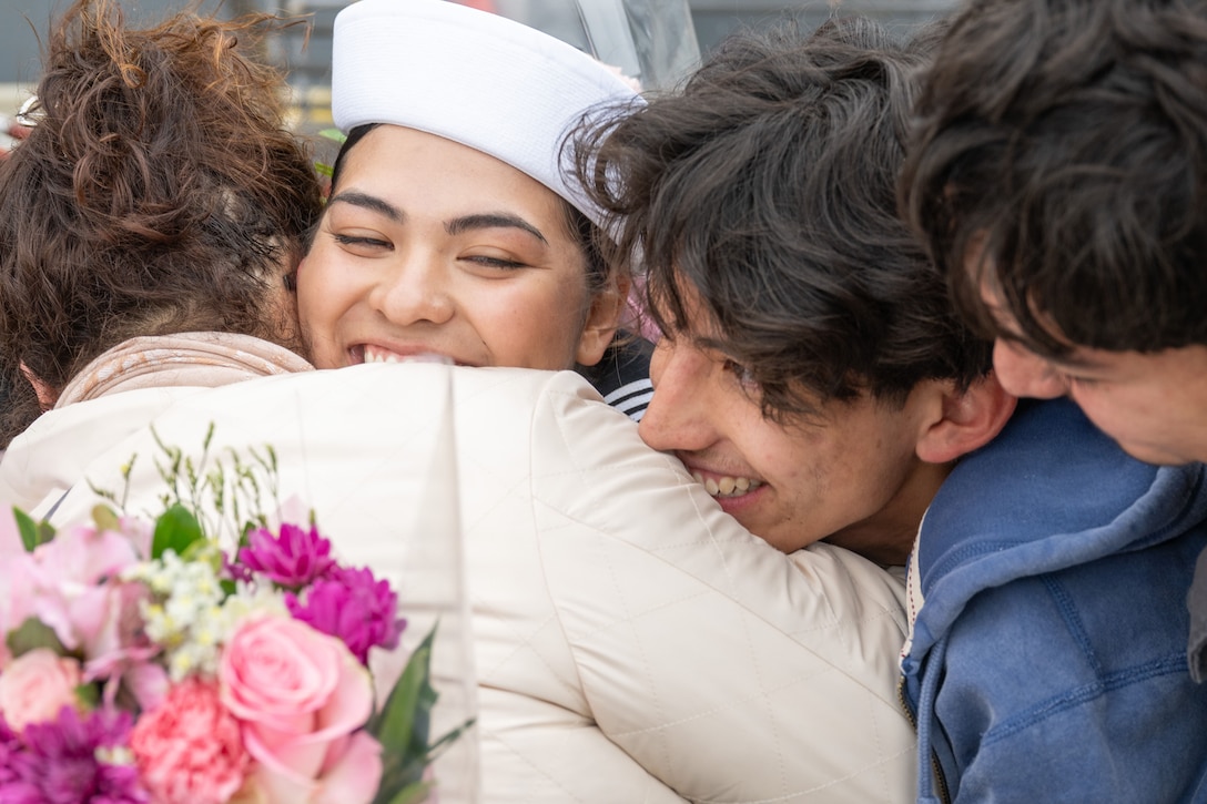 Family members holding flowers hug a smiling sailor.