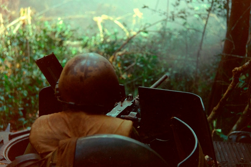 A man peeking out of the top of an armored vehicle fires a mounted gun into a jungle.