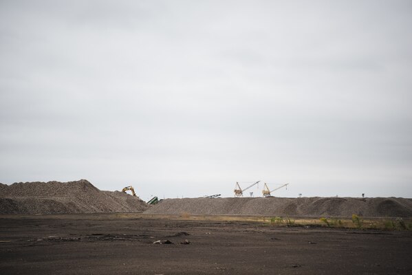 Undeveloped industrial property is visible in the foreground, with large piles of rock and gravel across the horizon. In the far background, cranes and industrial construction equipment is barely visible against the white, cloudy sky.