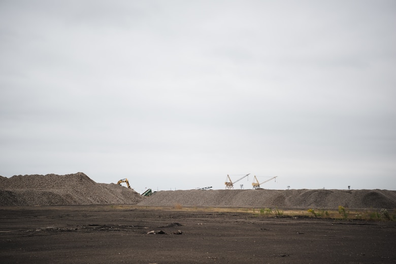 Undeveloped industrial property is visible in the foreground, with large piles of rock and gravel across the horizon. In the far background, cranes and industrial construction equipment is barely visible against the white, cloudy sky.