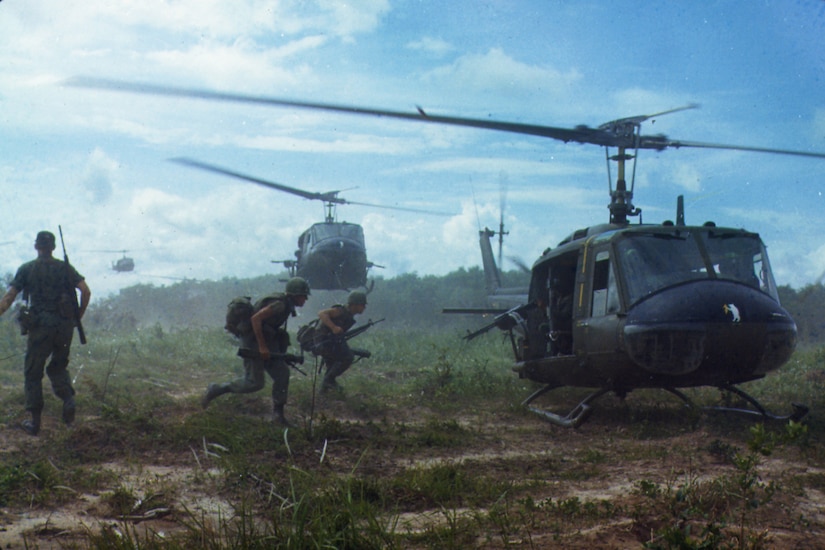 Soldiers run toward three helicopters landing in heavy brush during daytime.