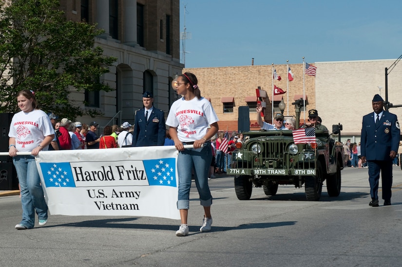 A man in the passenger seat of an old military vehicle waves during a parade in which two girls carry a banner in front of the vehicle that states the man's name.