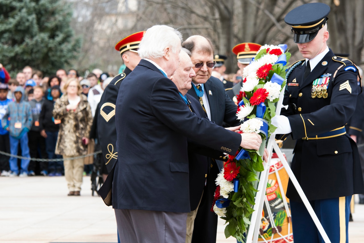 Four people prop up a large wreath on a mount as others look on from a distance.