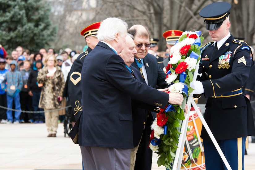 Four people prop up a large wreath on a mount as others look on from a distance.