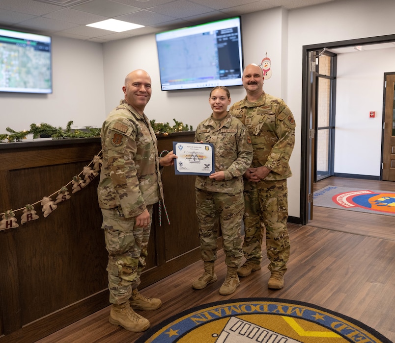U.S. Air Force Col. Adam Rosado, left, 97th Air Mobility Wing (AMW) deputy commander, and Chief Master Sgt. Jonny Adams, 97th AMW command chief, right, hand an award to Airman 1st Class Karissa Philyaw, 97th Operations Support Squadron airfield management operations coordinator, center, at Altus Air Force Base, Oklahoma, Dec. 17, 2025. Philyaw was named the Airman of the Month for November 2025. (U.S. Air Force Photo by Airman 1st Class Emma Wright) This photo has been altered for security purposes by blurring the two televisions.