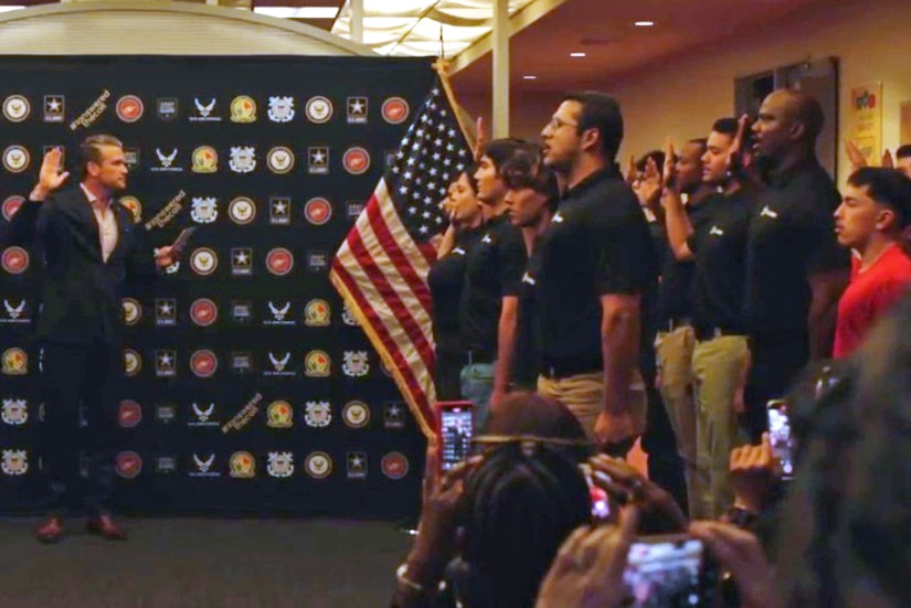 A person in civilian attire stands with an arm raised in front of military recruits, also with their right hands raised, indoors as other people look on; an American flag is also seen.