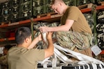 One Airman kneels on top of a container deliver system holding cargo netting while another stands beside the CDS taping the netting together