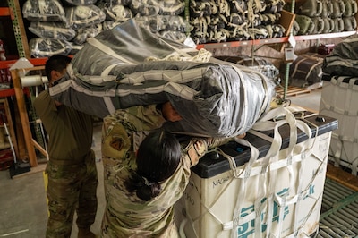 Two Airman heave a large packed parachute over their heads onto a container delivery system