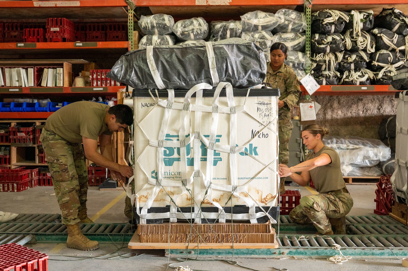 A container delivery system with a packed parachute on top sits in teh middle of the frame while Airmen on each side fasten strings to secure it and another stands at the back of the system