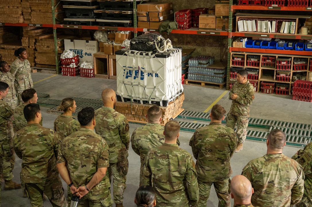 A half-circle of Airmen and Soldiers stand and listen to a colonel facing them