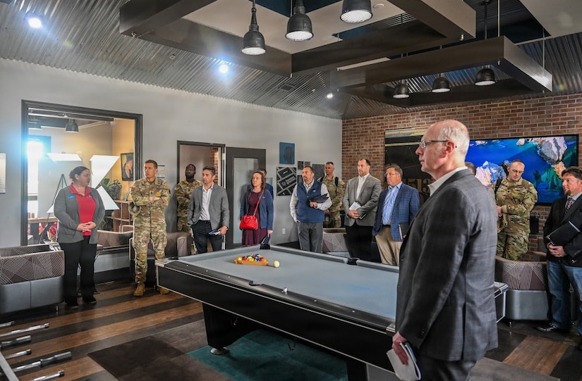 More than a dozen people, some in camouflage military uniforms and others in business attire, stand around a billiards table in a large room.