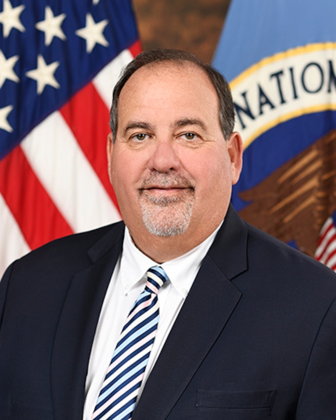 Photo of Timothy Kosiba, Deputy Director of the National Security Agency (NSA), standing in front of a flag of the United States and a flag showing the NSA seal.