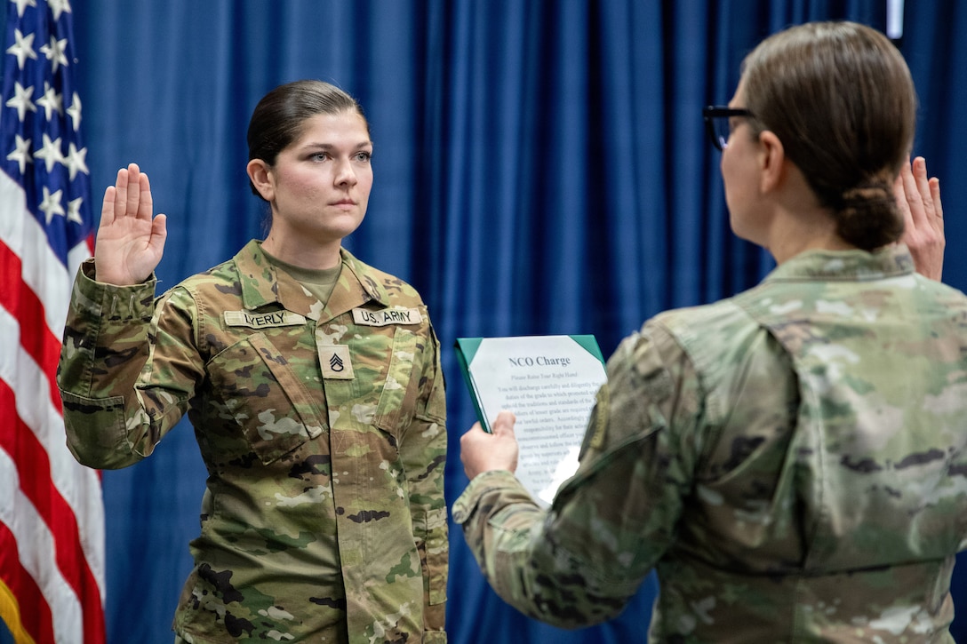 Two service members face each other with right hands raised, one holding a sheet of paper, while standing in front of blue curtains and an American flag.