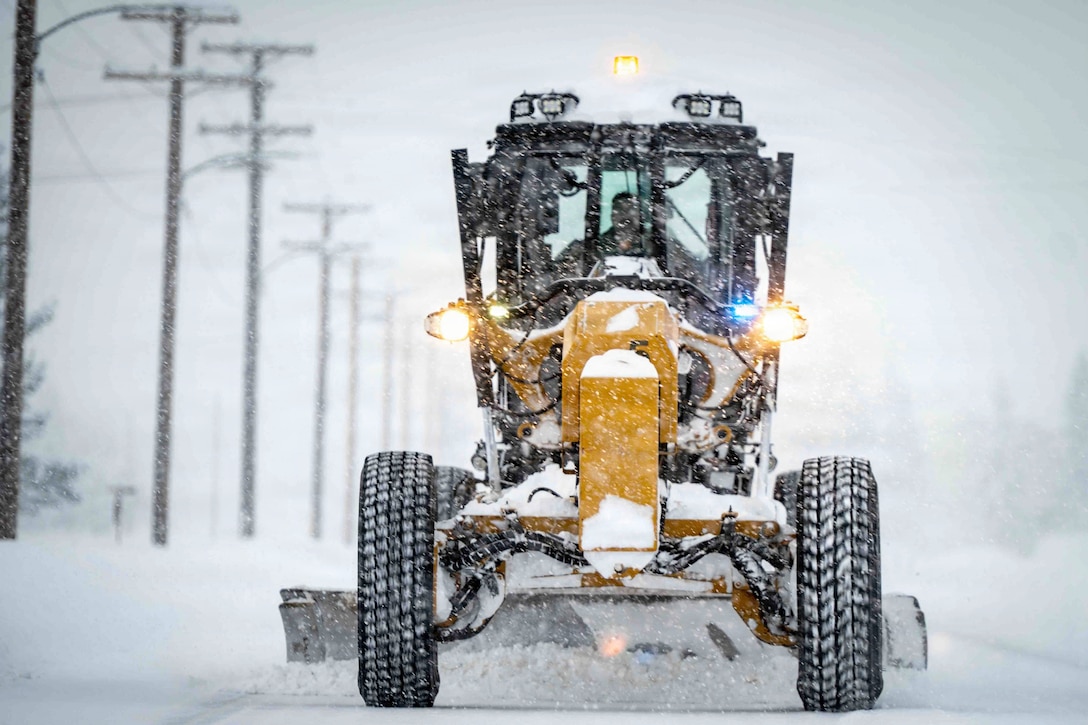 Air Force personnel operates a road grader in the snow illuminated by yellow lights.