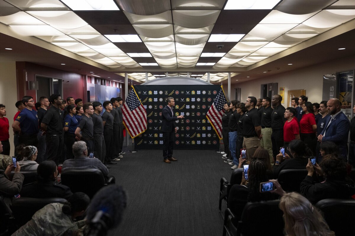 A man in business attire speaks to people in casual clothing, standing at attention, while others are seated, watching and taking photos.