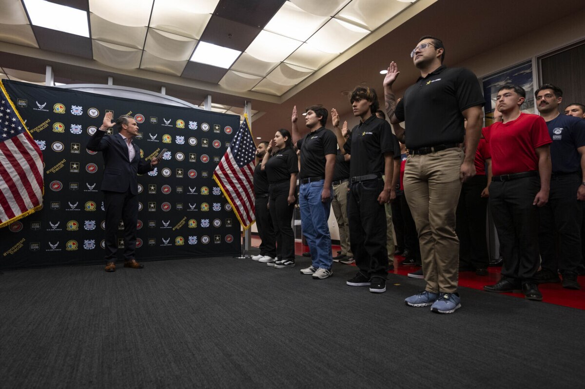 A man in business attire before an American flag, with his right hand raised, speaking to a group of people in casual clothing, all of them raising their right hands.