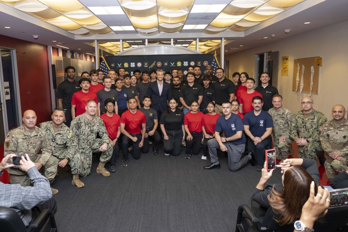 A large group of people in casual attire and camouflage military uniforms pose for a photo with a man in business attire at the center.