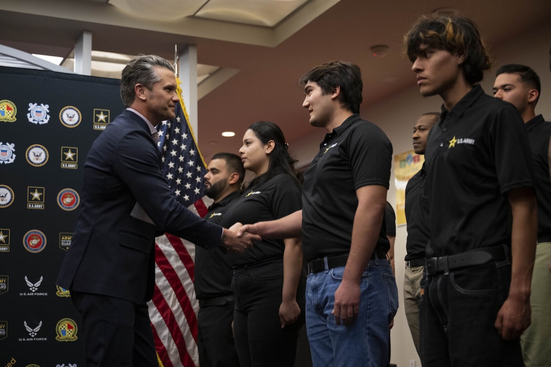 A man in business attire shakes hands with people in casual clothing in front of an American flag.