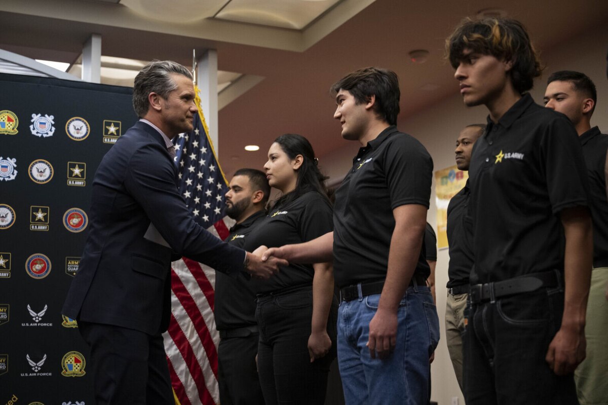 A man in business attire shakes hands with people in casual clothing in front of an American flag.