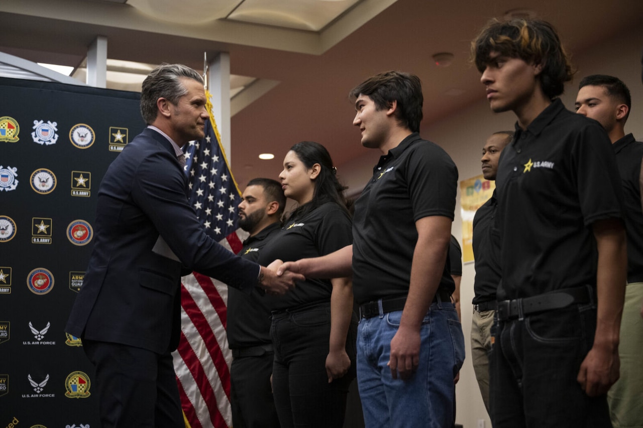 A man in business attire shakes hands with people in casual clothing in front of an American flag.