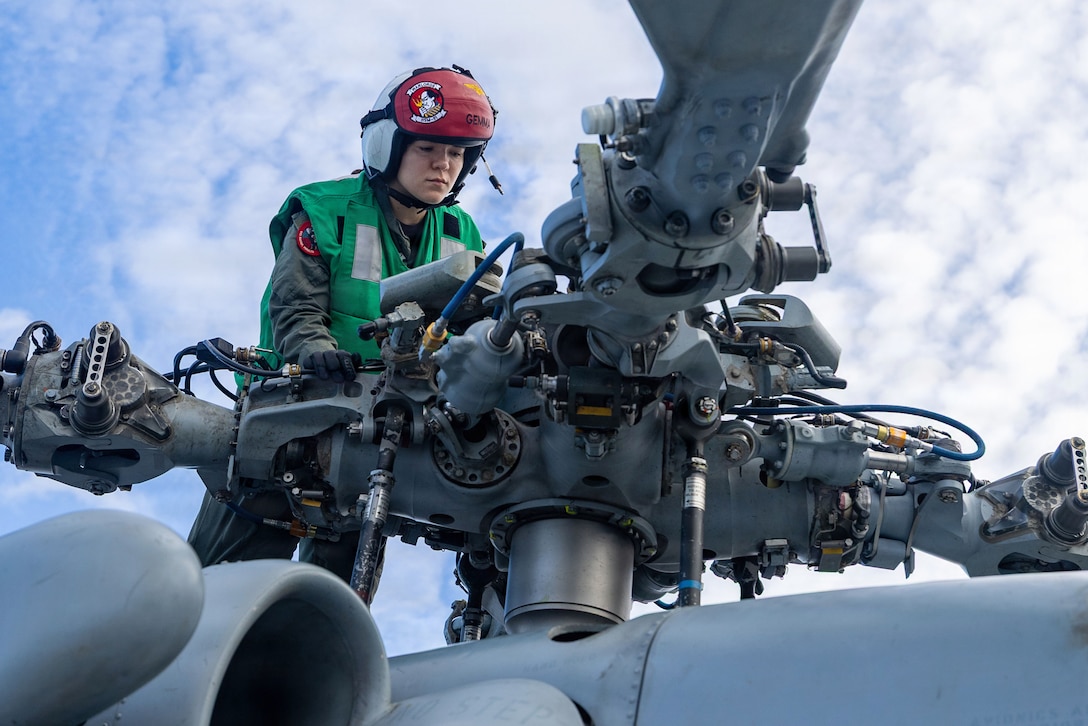 A sailor wearing protective gear inspects a helicopter's main rotor against a cloudy, blue sky.