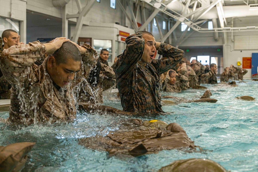 A large group of Marine Corps recruits shed gear in a pool while holding their hands on their heads.
