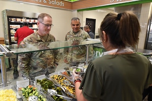 Two men dressed in military camouflage uniforms stand in front of a serving counter as a woman wearing an apron fills a bowl with ingredients for a salad.