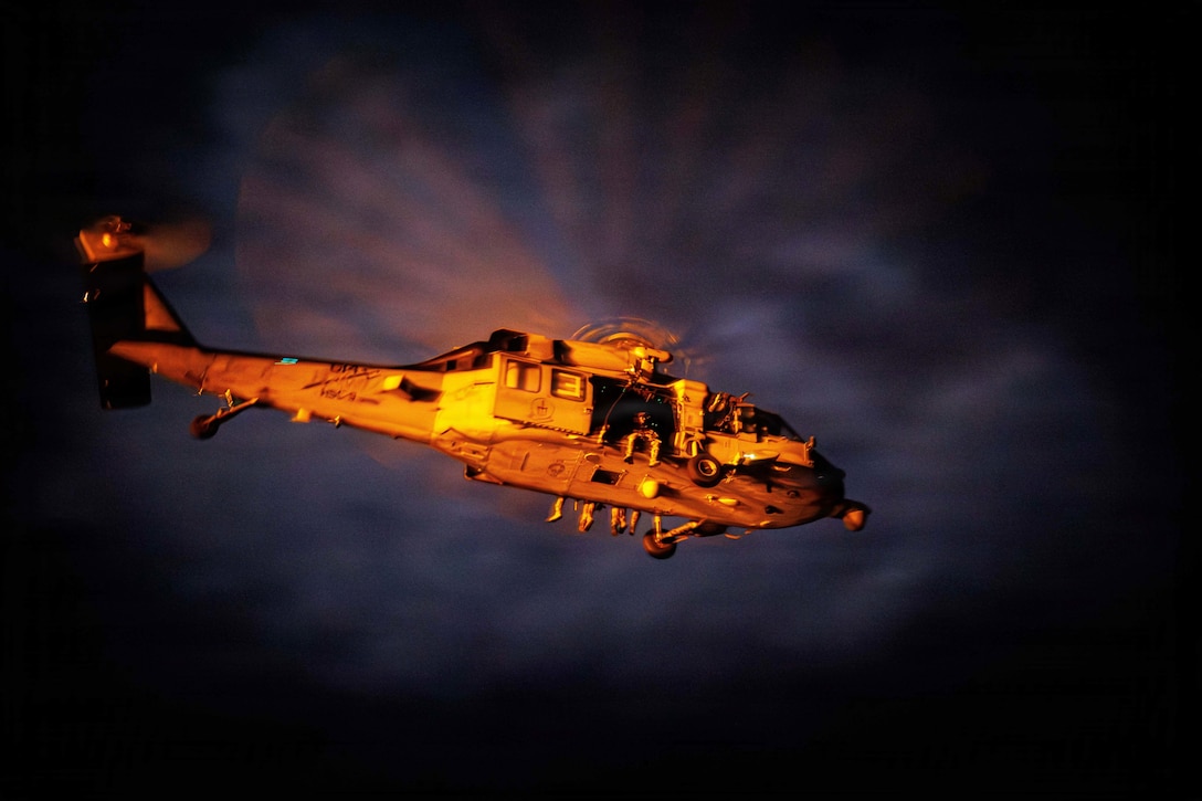 Service members sit in the open doorway of an airborne helicopter illuminated by a yellowish light.