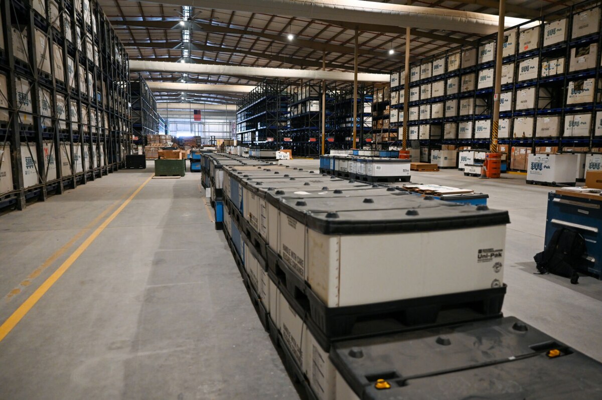 A military supply warehouse with rows of containers on the floor and similar containers stacked on shelves along the walls.