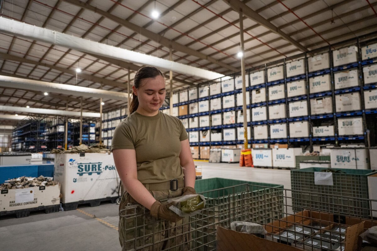 A woman in a camouflage military uniform holds a canteen and stands next to a wire basket inside a military supply warehouse. The walls of the building are lined with shelves holding boxes and containers.