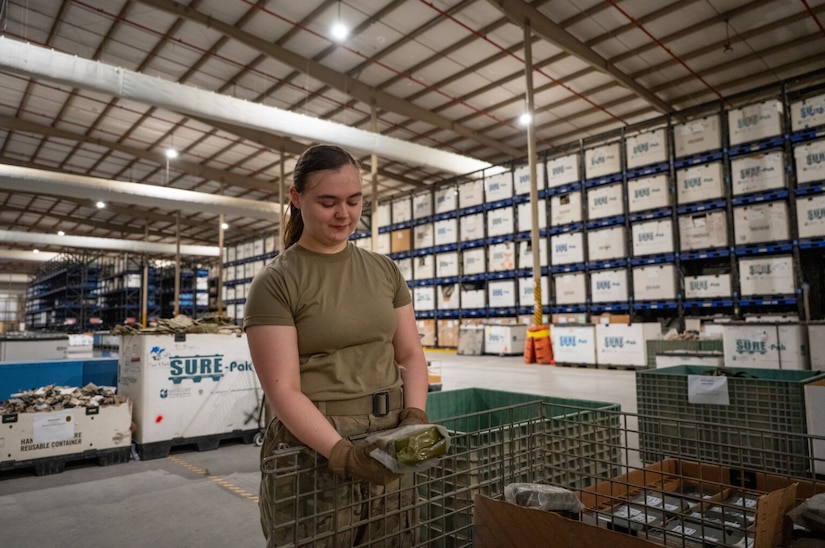 A woman in a camouflage military uniform holds a canteen and stands next to a wire basket inside a military supply warehouse. The walls of the building are lined with shelves holding boxes and containers.
