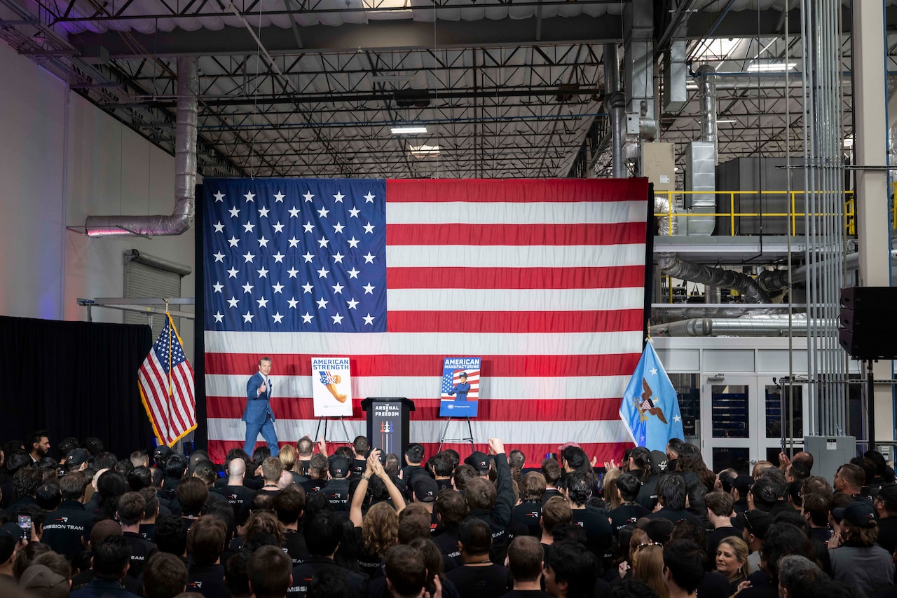 A man walks on a stage in front of an audience with the American flag in the background.